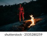 Hiker with backpack sits beside natural flames of Yanartaş, also known as eternal fire of Mount Chimaera, in Turkey. The night scene captures the unique fire phenomenon emerging from the mountain