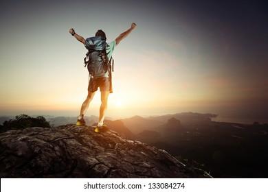 Hiker with backpack standing on top of a mountain with raised hands and enjoying sunrise