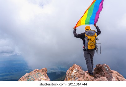 A Hiker With A Backpack Standing On The Top Of The Mountain And Holding A Pride Flag