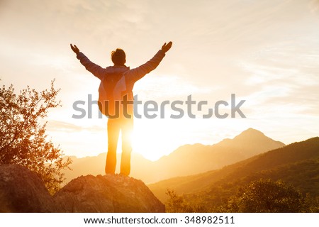 Hiker with backpack stands on che rock cliff in the mountains over the rising sun