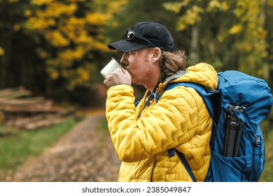 Hiker with backpack and yellow jacket drinking hot tea from travel mug in autumn forest. Refreshment and resting during outdoor hiking