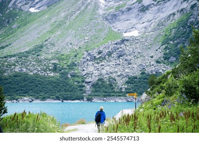 A hiker in a blue jacket stands by Gelmersee, surrounded by lush greenery and rugged, rocky slopes. The turquoise lake contrasts with the rocky mountain backdrop, creating a peaceful alpine scene. - Powered by Shutterstock