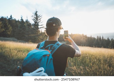 Hiker capturing the beauty of nature on a smartphone at sunset, standing in a grassy meadow with a forest backdrop. A blend of adventure, technology, and tranquility. - Powered by Shutterstock