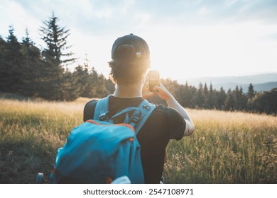 Hiker capturing the beauty of nature on a smartphone at sunset, standing in a grassy meadow with a forest backdrop. A blend of adventure, technology, and tranquility. - Powered by Shutterstock