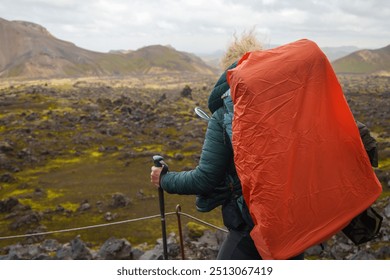 Hiker carrying a large orange backpack and trekking poles walking through the rugged volcanic terrain of Iceland, surrounded by rocky landscape and cloudy skies. - Powered by Shutterstock