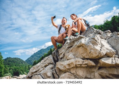 Hiker Couple With Small Yellow Dog Sitting On Mountain Peak And  Taking Picture With Smart Phone