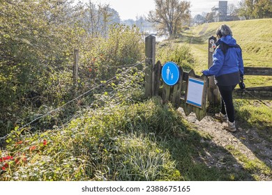 Hiker entering Belgian nature reserve De Wissen Maasvallei, round plate with sign pedestrian route or walking area at gate, river and trees in misty background, sunny day in Dilsen-Stokkem, Belgium - Powered by Shutterstock