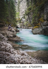 Hiker In Flannel Shirt And Hat Sitting Along Maligne River In Maligne Canyon, Jasper National Park, Alberta, Canada. Long Exposure, Cyan, Turquoise, Swift, Wild River, Valley. Magical, Calm, Peaceful.