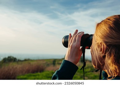 A hiker girl looks through binoculars at nature and birds standing on the top of a mountain in the bright rays of setting sun against the background of beautiful spring nature and cloudy blue sky. - Powered by Shutterstock