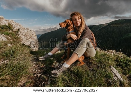Hiker girl sitting with pointer dog at mountain top and enjoying views of coniferous forest of mountain range Kopaonik. Woman with Hungarian Vizsla, trekking and travel with pets lifestyle concept.