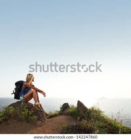 Hiker lady with backpack relaxing on a rock by sea and looking to the horizon