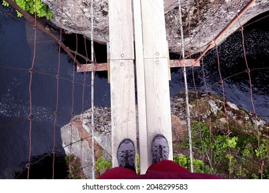 Hiker Looking Down At Feet At River Below On A Rope Bridge In Norway