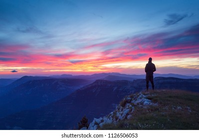 A hiker is looking at a tranquil, pink sunset in a mountainous wilderness.