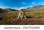 Hiker with Lopapeysa nordic sweater and red beannie enjoys spectacular scenery to the mountain valley with dramatic tall peaks and sunny grassy terrain of arctic wilderness on Kungsleden trail, Sweden