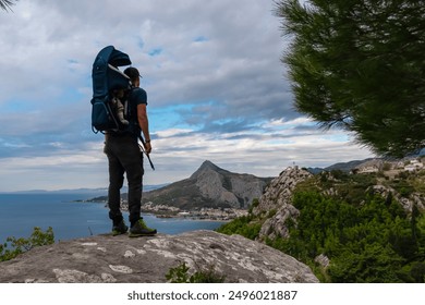 Hiker man with baby carrier backpack on rock formation with aerial view of coastal town Omis, Dinara mountains, Split-Dalmatia, Croatia, Europe. Coastline of Adriatic Sea, Balkans. Travel destination - Powered by Shutterstock