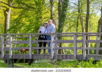 Hiker mature couple smiling and happy on a wooden bridge with their dachshund, looking at the camera, lush green trees in the background, sunny spring day in Stein, South Limburg, the Netherlands - Powered by Shutterstock