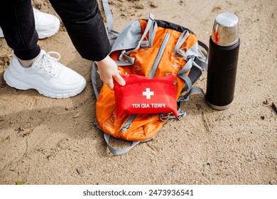 A hiker packs essentials in an orange daypack with a red first aid kit and thermos on a sandy beach, preparing for a wilderness adventure. Safety gear for outdoor activities and travel - Powered by Shutterstock