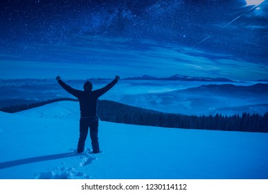 Hiker with raised hands standing on a snowy hill at night. Milky way in a starry sky above the mountain valley.
