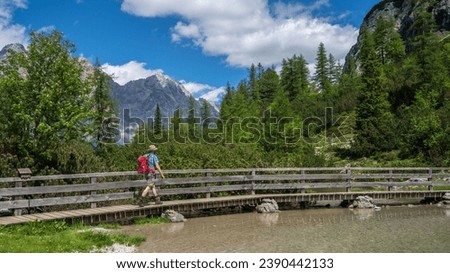 A hiker with a red backpack is walking over a wooden bridge. in the background are the Wetterstein mountains.