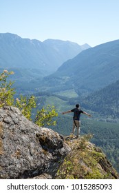 Hiker Standing With Arms Wide Open, On The Edge Of A Mountaintop Overlooking A Valley, With A Vista Of Trees And Hills On The Hazy Horizon 