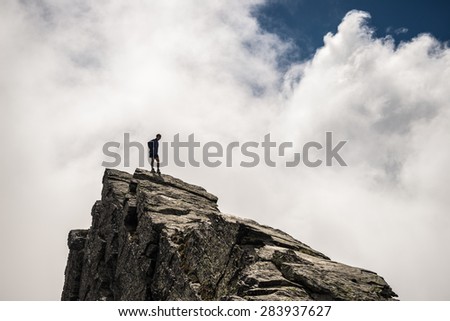 Hiker standing on steep rocky mountain summit without protection. Dramatic cloudy sky. Concept of reaching the goal and conquering the success. Contrasted, silhouette like.