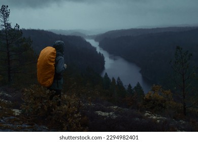 Hiker standing on top of a hill in a forest looking out over a river in rainy weather wearing a backpack.