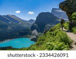 A hiker standing on a trail under a rock overhang watching a scene of a lake below and mountain peaks above, Grinnell Glacier Trail, Glacier National Park, Montana