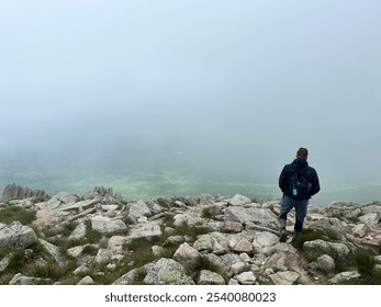 A hiker stands on a rocky mountain peak shrouded in mist, gazing into the foggy landscape below, creating a sense of solitude and awe. - Powered by Shutterstock