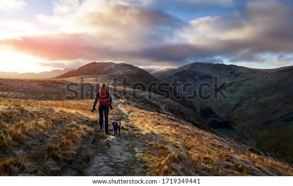 A hiker and their dog walking towards the\
mountain summit of High Spy from Maiden Moor at sunrise on the\
Derwent Fells in the Lake District,\
UK.