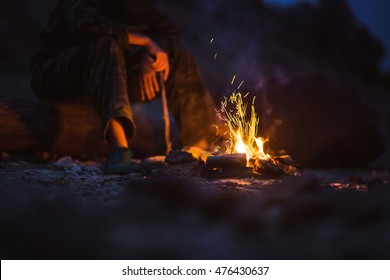 Hiker Warms Their Feet Next To A Campfire At Dusk Camping In The Woods