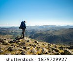 A hiker wearing a backpack and carrying trekking poles views the horizon from Great Gable, a fell (or mountain) in the Lake District, Cumbria, England.