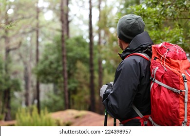 Hiker wearing hiking backpack and hardshell jacket on hike in forest. Man wearing hat gloves using hiking sticks poles outdoors in woods. Male hiker standing looking away. - Powered by Shutterstock