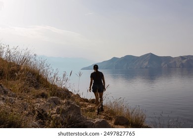 Hiker woman on coastal hiking trail along rocky cliffs overlooking calm Adriatic sea near Baska, Krk Otok, Primorje-Gorski Kotar, Croatia. Majestic mountain range emerges from abandoned island Prvic - Powered by Shutterstock