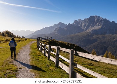 Hiker woman on scenic hiking trail along wooden fence on alpine meadow. View of massive mountain ridges of majestic untamed Sexten Dolomites in South Tyrol, Italy, Europe. Hiking concept Italian Alps