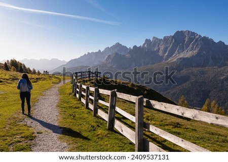 Hiker woman on scenic hiking trail along wooden fence on alpine meadow. View of massive mountain ridges of majestic untamed Sexten Dolomites in South Tyrol, Italy, Europe. Hiking concept Italian Alps