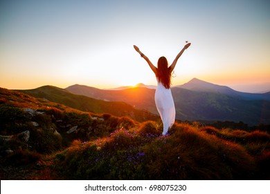 Hiker woman standing with hands up achieving the top. Girl welcomes a sun. Conceptual design. Successful woman hiker open arms on sunrise mountain top. Girl in long white dress in the mountains.