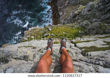 Hiker's legs hanging over the edge of a cliff with the blue ocean far beneath.