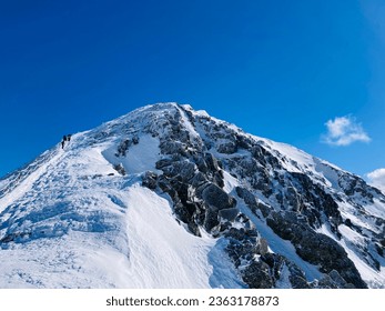 Hikers and mountaineers summiting the western side of Sněžka peak in Krkonoše, Czech Republic. Winter hiking trail from low angle with unrecognizable figures in the distance. Snowy hill with blue sky. - Shutterstock ID 2363178873