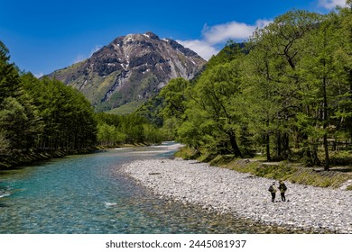 Hikers on the bank of a clear river leading towards snow-capped mountains (Japanese Alps, Nagano) - Powered by Shutterstock