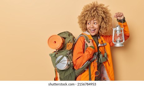 Hiking and adventure concept. Positive active female camper explores new places holds lantern to see in darkness carries backpack isolated over brown background empty space for your promotion