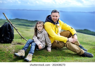 Hiking Family Adventure On Mountain Trek: Beauty Kid Girl And Her  Active Father In Jackets And Special Boots Having Rest On Green Lawn With Mountain Lake View: Scotland, Skye