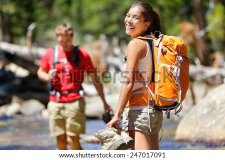 Hiking friends having fun crossing river in forest. Young happy adults barefoot walking in water with wet feet on an adventure trip hike in nature.