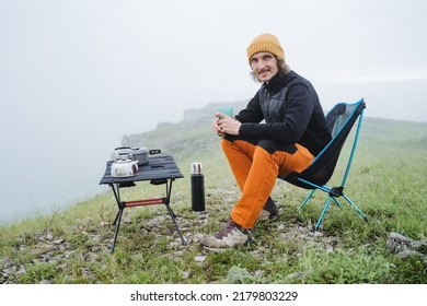 Hiking In The Mountains Camping Camping Sitting On A Chair, A Guy Sitting Smiling Holding A Glass Of Tea, A Folding Table For Eating In Nature, A Climber Has Breakfast High On A Rock