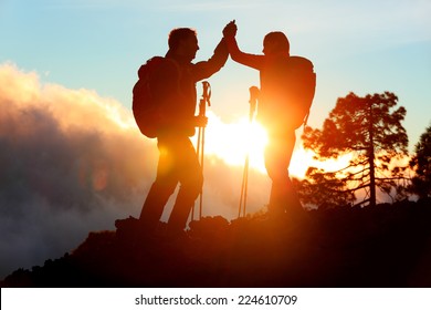 Hiking people reaching summit top giving high five at mountain top at sunset. Happy hiker couple silhouette. Success, achievement and accomplishment people