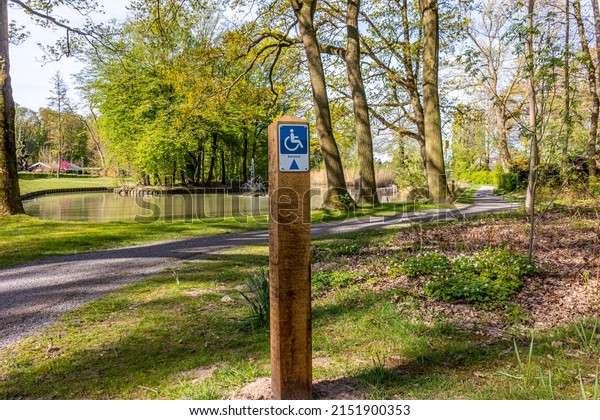 Hiking sign for disabled people in a park,\
drawing and the word Rolstoel means wheelchair, a path, pond and\
trees in the background, sunny day in Heidekamp Park, Stein, South\
Limburg, Netherlands