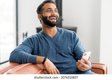 Hilarious 30s Indian Man Using Smartphone Sitting On The Couch At Home, Hispanic Latin Male Holds Mobile Phone And Laughing Cheerfully, Watching Comedy, Funny Videos, Joking With Friends Online