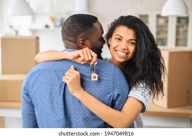 Hilarious biracial woman embraces black man and holds keys with trinket in form of cute wooden house, looks happily at the camera. African-American couple bought own property, moved in new apartment