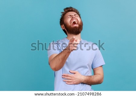 Hilarious laughter. Portrait of joyful happy bearded man laughing loudly and pointing to camera, mocking taunting you, holding belly. Indoor studio shot isolated on blue background.