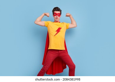 Hilarious male in bright superhero cape and mask demonstrating strength and power while showing muscles on blue background in studio and looking at camera