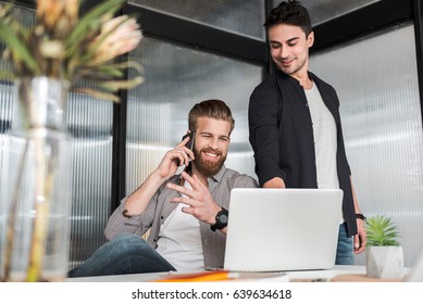 Hilarious manager is standing beside his co-worker, sitting near table and speaking on phone. Men are looking at screen of laptop